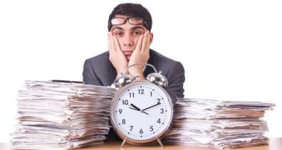 Harried-looking man sitting behind a clock and two large stacks of paper