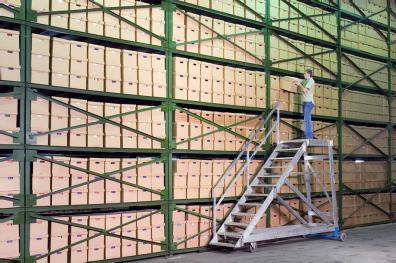 Man standing on rolling staircase selecting a box from a tall shelf in a warehouse