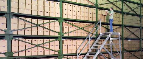 Man standing on rolling staircase selecting a box from a tall shelf in a warehouse