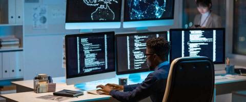 Cybersecurity specialist sits at a desk with five screens monitoring threat levels. 