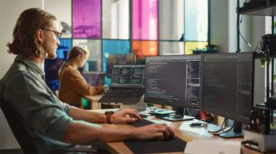 An IT Support professional working intently at a desk with multiple monitors displaying complex code, in an office with a colleague in the background.