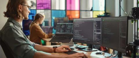 An IT Support professional working intently at a desk with multiple monitors displaying complex code, in an office with a colleague in the background.